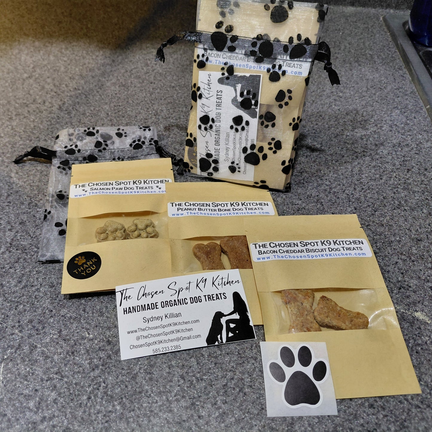 3 Dog treat packages layed out diagonally, Salmon Paws, Peanut Butter Bones & Bacon Cheddar Biscuits with visible branding for The Chosen Spot K9 Kitchen