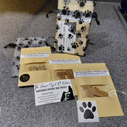 3 Dog treat packages layed out diagonally, Salmon Paws, Peanut Butter Bones & Bacon Cheddar Biscuits with visible branding for The Chosen Spot K9 Kitchen