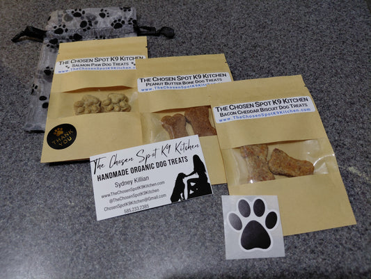 A mesh bag with paw-prints, 3 Dog treat packages layed out diagonally, Salmon Paws, Peanut Butter Bones & Bacon Cheddar Biscuits with visible branding for The Chosen Spot K9 Kitchen and a paw-print sticker.