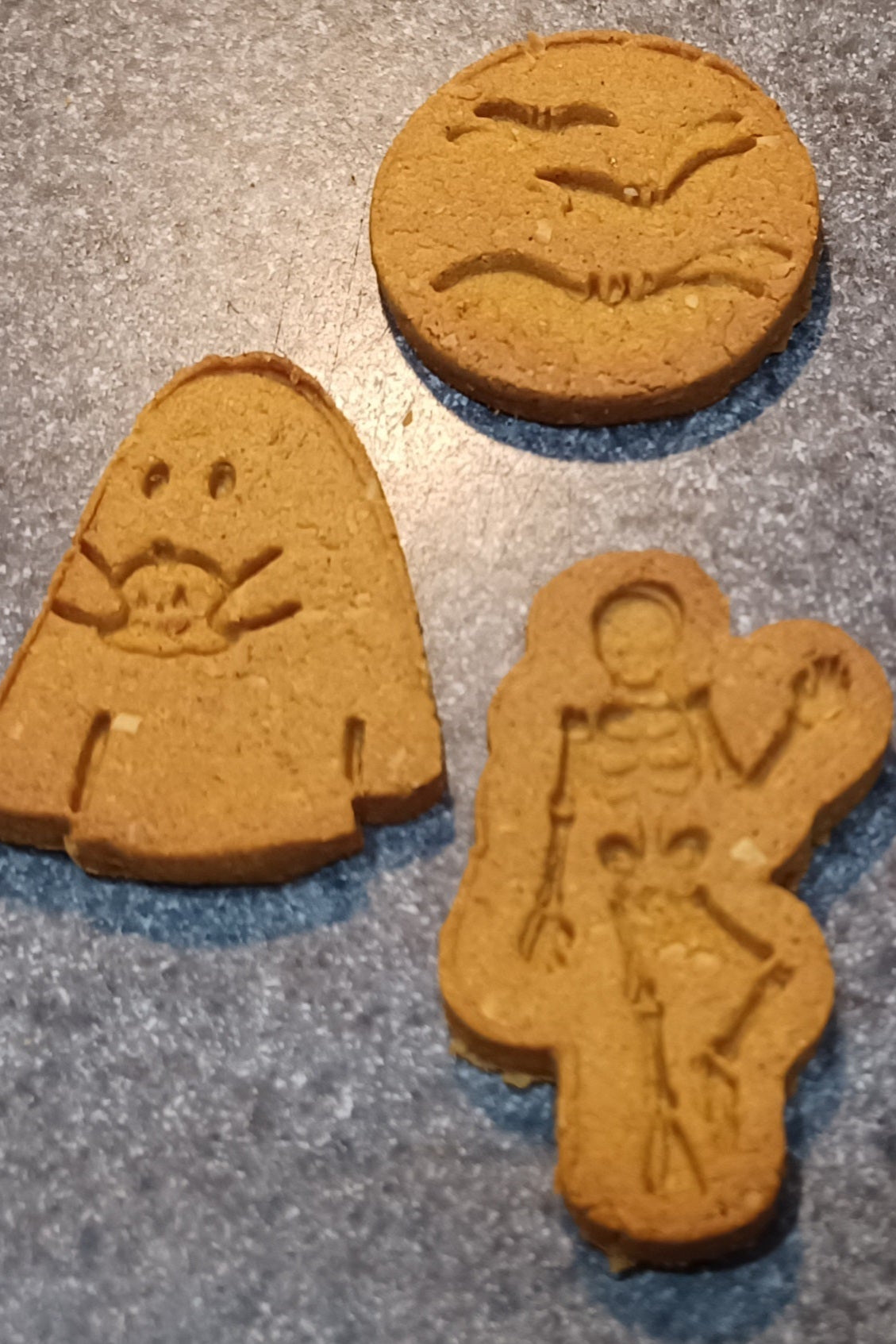 Three cookies shaped like a skeleton, a ghost, and a moon with bats on a gray surface.
