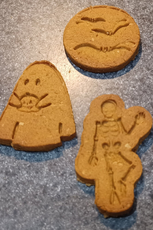 Three cookies shaped like a skeleton, a ghost, and a moon with bats on a gray surface.